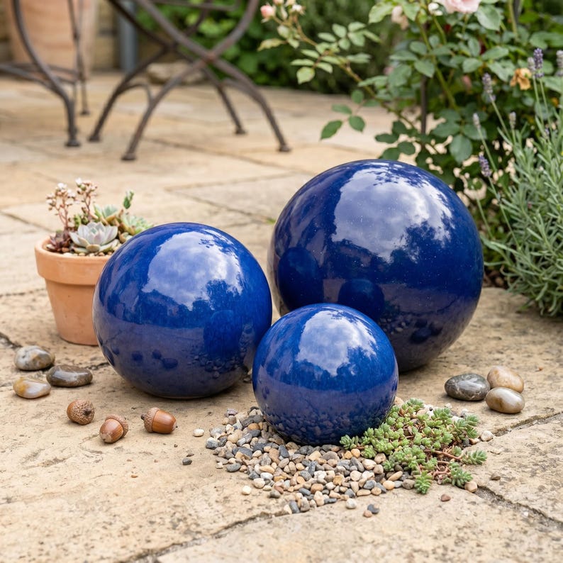 May include: Three glossy, deep blue ceramic spheres of varying sizes are arranged on a stone patio. The spheres reflect the sky and surrounding greenery. Small pebbles and greenery surround the spheres, with a terracotta pot and acorns in the background.