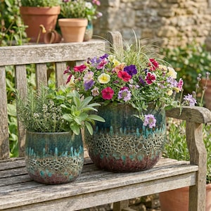May include: Two ceramic planters with colorful flowers and herbs sit on a wooden bench. The planters have a textured, teal and brown glaze. One planter is filled with vibrant petunias, while the other contains herbs.