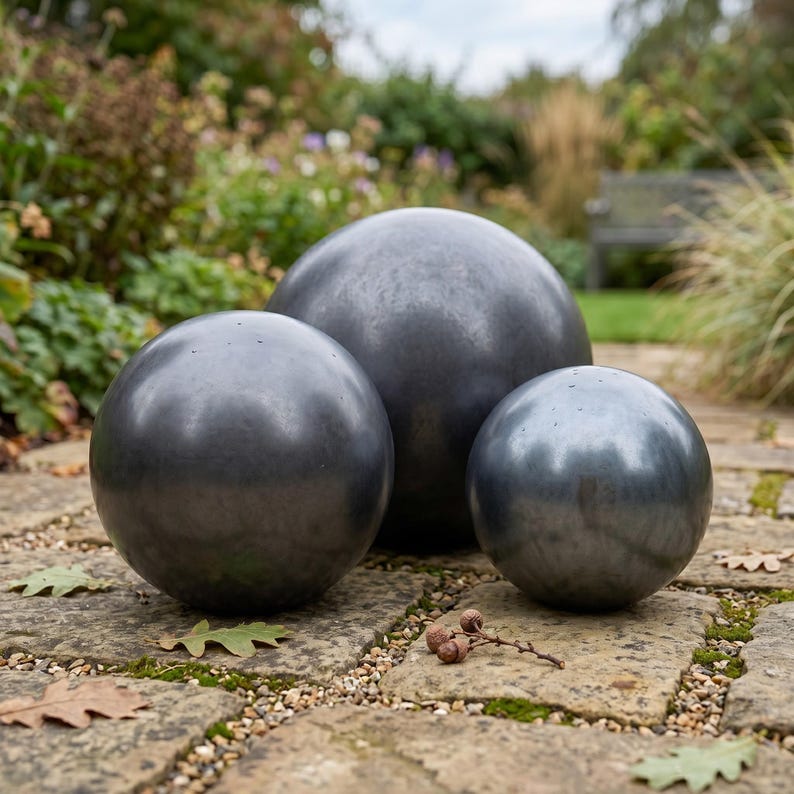 May include: Three dark gray metal spheres of varying sizes are arranged on a stone surface. The spheres have a smooth, reflective surface. The background features greenery and a glimpse of a bench, suggesting an outdoor garden setting.