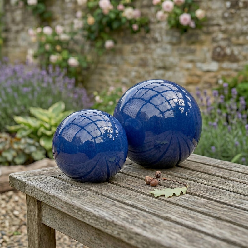 May include: Two glossy, deep blue decorative spheres of different sizes sit on a weathered wooden table. The spheres reflect the surrounding garden, including a stone wall and flowering plants. Acorns and a leaf rest on the table.