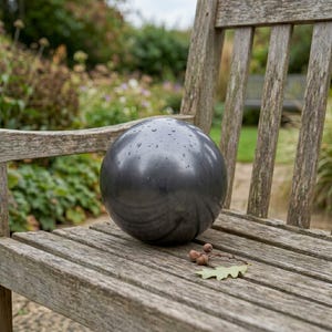 May include: A dark gray, spherical garden ornament with water droplets on its surface. The ball is resting on a weathered wooden bench, with a natural outdoor background. A few acorns and a leaf are also on the bench.
