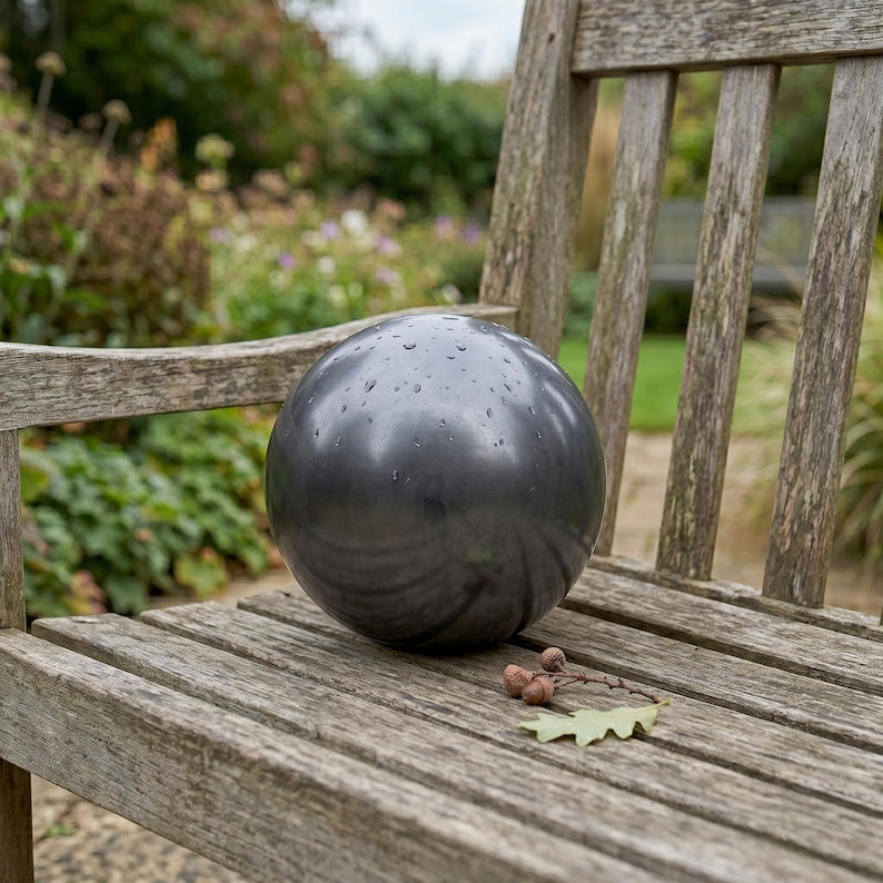 May include: A dark gray, spherical garden ornament with water droplets on its surface. The ball is resting on a weathered wooden bench, with a natural outdoor background. A few acorns and a leaf are also on the bench.