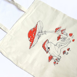 May include: A white canvas tote bag with a black and red illustration of a frog sitting on a mushroom. The mushroom has red and white spots and there are other mushrooms around the frog.