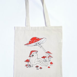 May include: A white canvas tote bag with a red and white mushroom design. A frog sits under the mushroom with its legs outstretched. There are smaller mushrooms surrounding the frog.