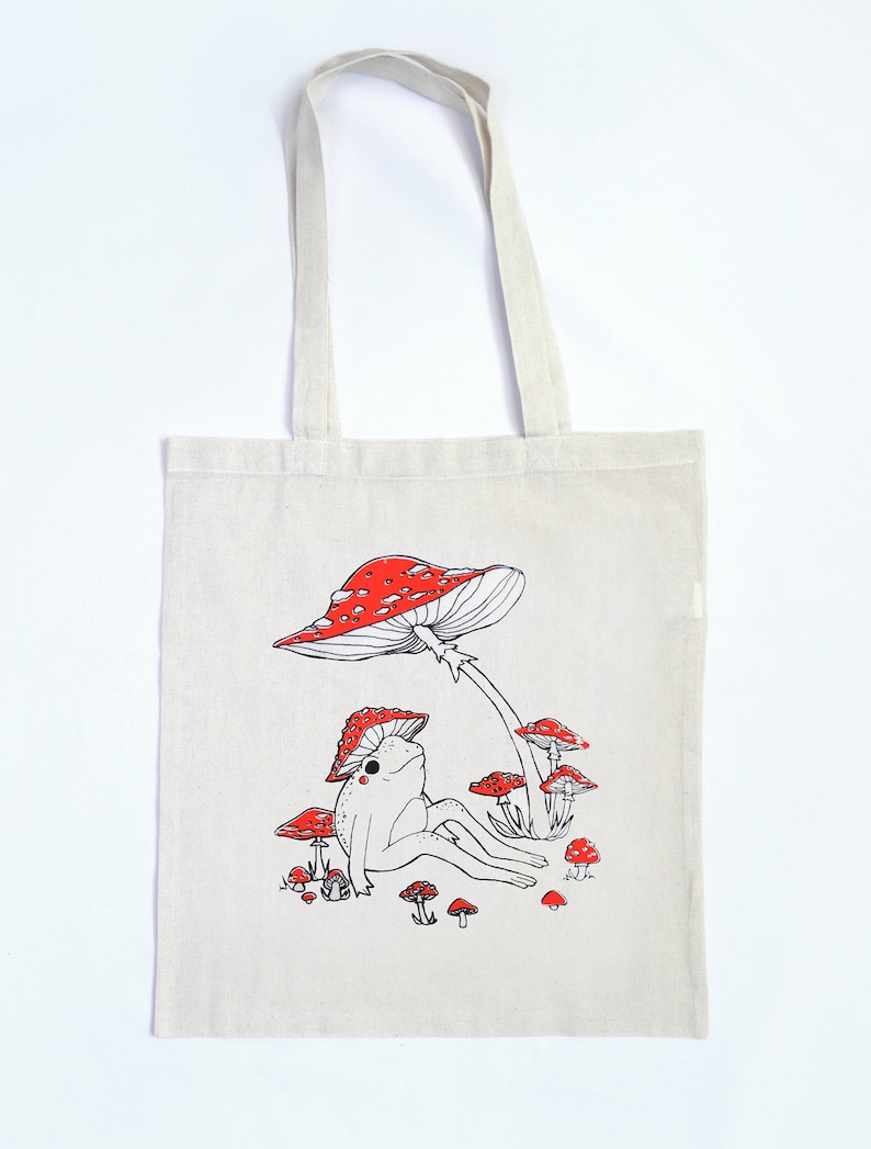 May include: A white canvas tote bag with a red and white mushroom design. A frog sits under the mushroom with its legs outstretched. There are smaller mushrooms surrounding the frog.