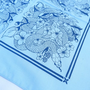 Puede incluir: Un pañuelo azul claro con un diseño en blanco y negro de una serpiente y flores. El pañuelo tiene un borde azul oscuro.