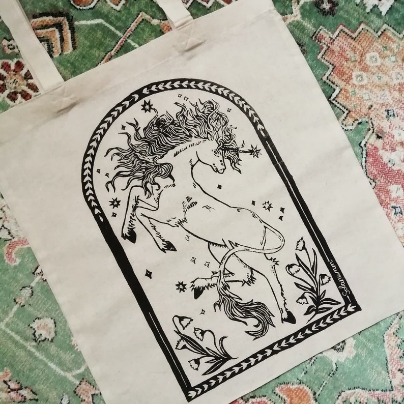 May include: A black and white tote bag featuring a graphic of a unicorn with a star on its horn. The unicorn is surrounded by stars and flowers. The text "Sojourn" is printed on the bag.
