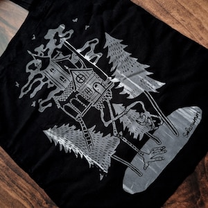 May include: Black and white tote bag featuring a graphic design of a house on stilts in a forest. The house has a window and a door. The trees are stylised and have a whimsical feel. The text "Solarumin" is printed in the bottom right corner of the bag.