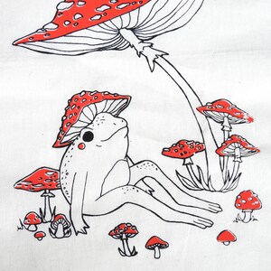 May include: A black and white line drawing of a frog sitting under a red and white mushroom. The frog is surrounded by smaller red and white mushrooms.