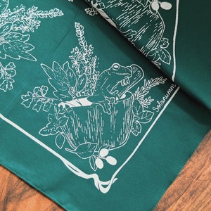 Könnte beinhalten: Ein grünes Bandana mit einem weißen Design eines Frosches, der in einem Topf mit Pflanzen sitzt. Das Bandana trägt den Schriftzug "Solanum".