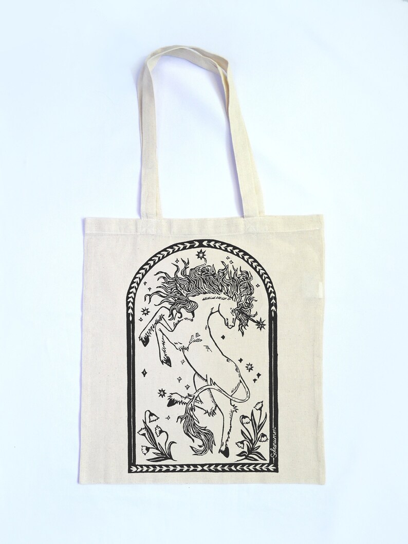 May include: A white tote bag with black print of a unicorn in a star-filled archway with flowers. The text "Seleneum" is at the bottom of the design.