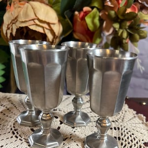 Peut inclure: Quatre gobelets vintage en étain avec un design à facettes. Les gobelets ont une tige et une base classiques, et sont disposés en groupe. L'arrière-plan comprend des fleurs et un napperon en dentelle.
