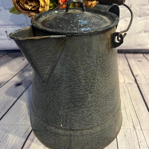 Könnte beinhalten: Eine vintage graue Emaillekanne mit einem Holzgriff und einem Deckel. Die Kanne hat eine gesprenkelte Oberfläche und einen Ausguss an der Seite.