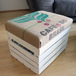 Könnte beinhalten: Weißer Holzkasten mit einer Kissenauflage aus Sackleinen. Das Kissen hat ein grünes Blattdesign und den Text "Café do Brasil".