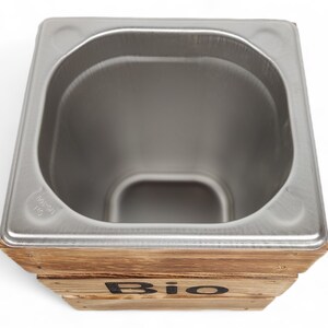 May include: A stainless steel compost bin with a wooden frame. The bin has a rounded top and the word "Bio" is printed on the front of the frame.