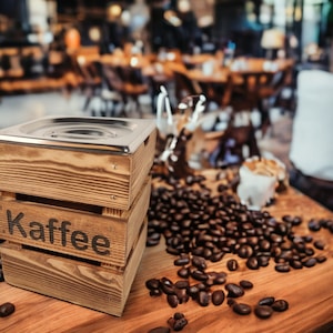 May include: A wooden box with a metal lid, labeled "Kaffee", sits on a table surrounded by coffee beans.