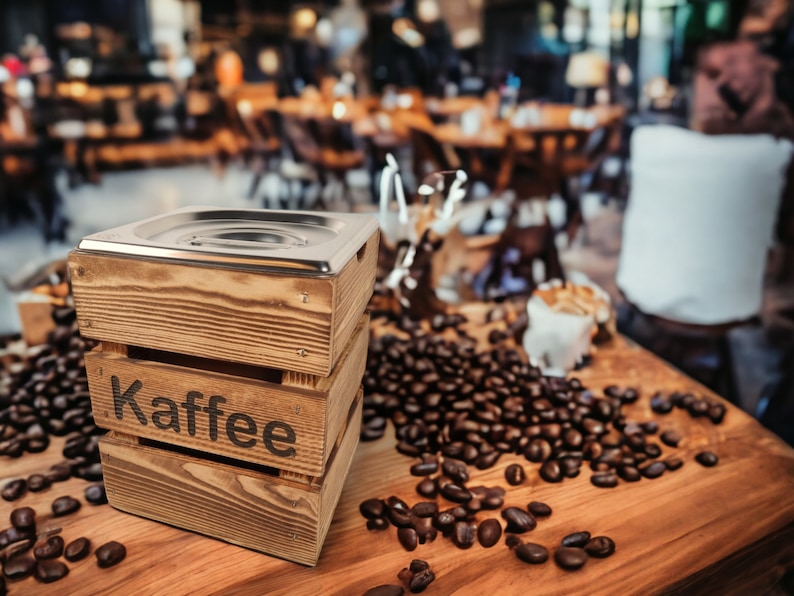 May include: A wooden box with a metal lid, labeled "Kaffee", sits on a table surrounded by coffee beans.