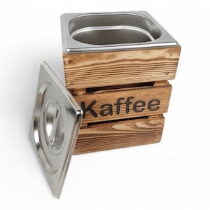 May include: A wooden box with a stainless steel lid and the word "Kaffee" printed on the front. The box is made of natural wood with a rustic finish.