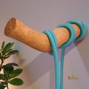 May include: A wooden coat rack fixed to a wall, with a curved, natural wood design. The rack is wrapped with teal-coloured rope. The design is simple and modern, suitable for a minimalist aesthetic. The rack is approximately 20 cm long.