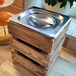 May include: A wooden compost bin with a stainless steel lid. The bin is made of natural wood and has a rustic look. The lid has a handle for easy opening and closing.