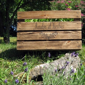 May include: A wooden crate with the words "Jardin d'herbes" burned into the wood. The crate is sitting on a rock with lavender plants growing around it.
