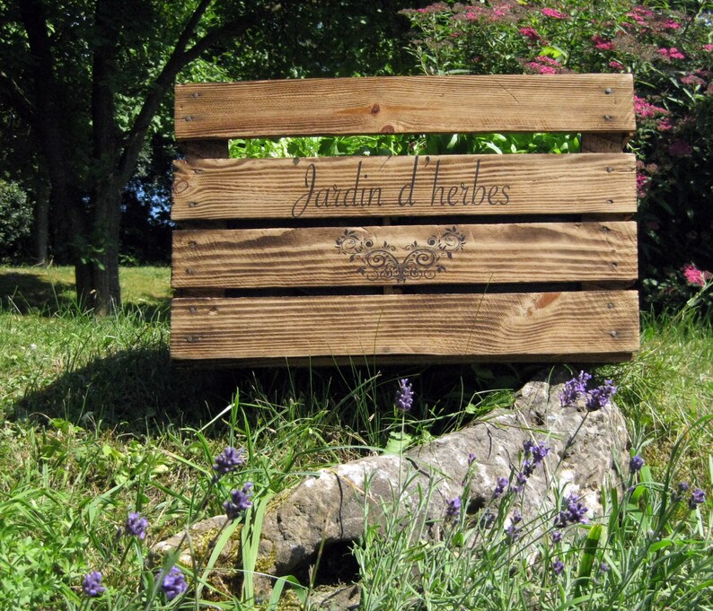 May include: A wooden crate with the words "Jardin d'herbes" burned into the wood. The crate is sitting on a rock with lavender plants growing around it.