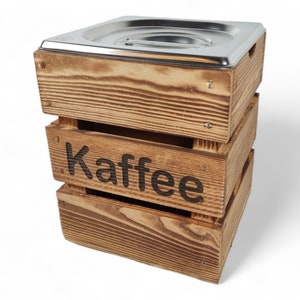 May include: A wooden box with a metal lid, stacked on top of two other wooden boxes. The box is labeled "Kaffee" in black lettering.