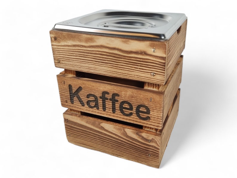 May include: A wooden box with a metal lid, stacked on top of two other wooden boxes. The box is labeled "Kaffee" in black lettering.