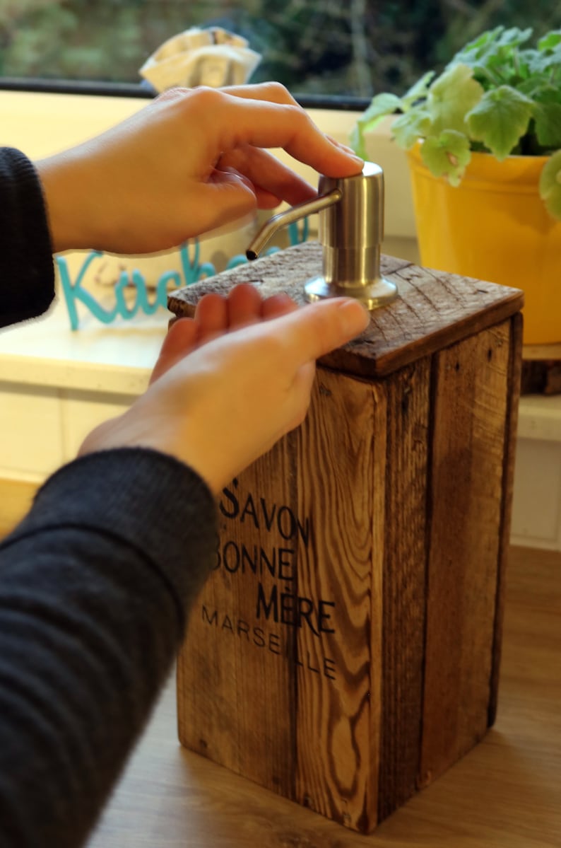 May include: A wooden soap dispenser with a stainless steel pump. The dispenser is labeled "Savon Bonne Mere Marseille".