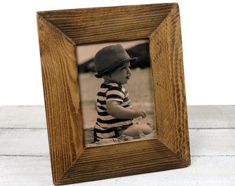 Può includere: Una cornice per foto in legno rustico con una foto di un bambino che indossa un cappello e una camicia a righe. La cornice è di colore marrone scuro e ha una finitura invecchiata.