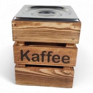 May include: A wooden box with a metal lid, labeled "Kaffee" in black lettering. The box is made of light brown wood with a rustic finish.