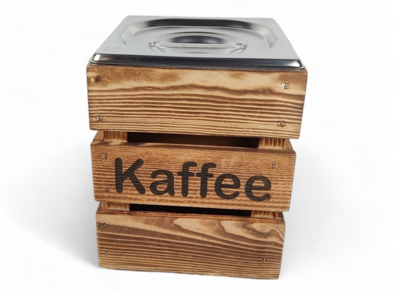 May include: A wooden box with a metal lid, labeled "Kaffee" in black lettering. The box is made of light brown wood with a rustic finish.