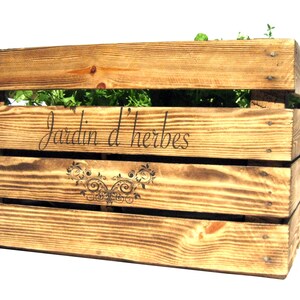 May include: A wooden crate planter with the text "Jardin d'herbes" and a decorative design. The planter is filled with green herbs.