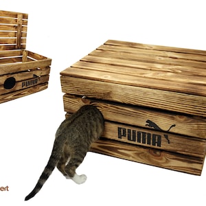 May include: A wooden cat house with a hinged lid and a Puma logo. The house is made of natural wood with a rustic finish. A cat is entering the house through a hole in the front.
