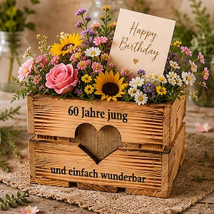 Puede incluir: Una caja de madera rústica con un recorte en forma de corazón, llena de un colorido arreglo floral de rosas rosadas, girasoles amarillos y pequeñas margaritas. Una tarjeta con el texto Happy Birthday está insertada. La caja tiene el texto alemán 60 Jahre jung und einfach wunderbar.
