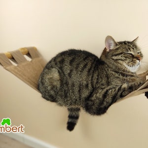 May include: A tabby cat relaxes on a wall-mounted cat hammock. The hammock is made of tan fabric and wooden supports. The cat is brown and black with its eyes closed, enjoying the comfort of its new bed.
