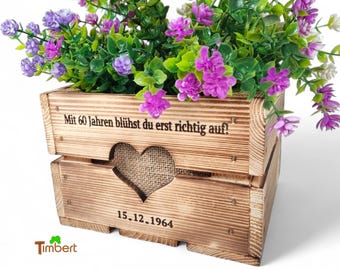 May include: A rectangular wooden crate with a burnt finish, featuring a heart-shaped cutout and burlap backing. The text "Mit 60 Jahren blühst du erst richtig auf!" and "15.12.1964" are engraved on the front. The crate is filled with purple and white artificial flowers.