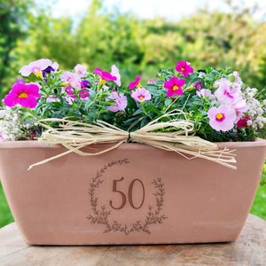 Puede incluir: Una maceta de terracota con una corona decorativa y el número 50 grabado en ella. La maceta está llena de flores rosas y blancas.