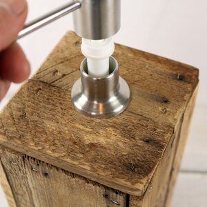 May include: A wooden soap dispenser with a stainless steel pump. The pump has a white plastic ring and a silver handle.