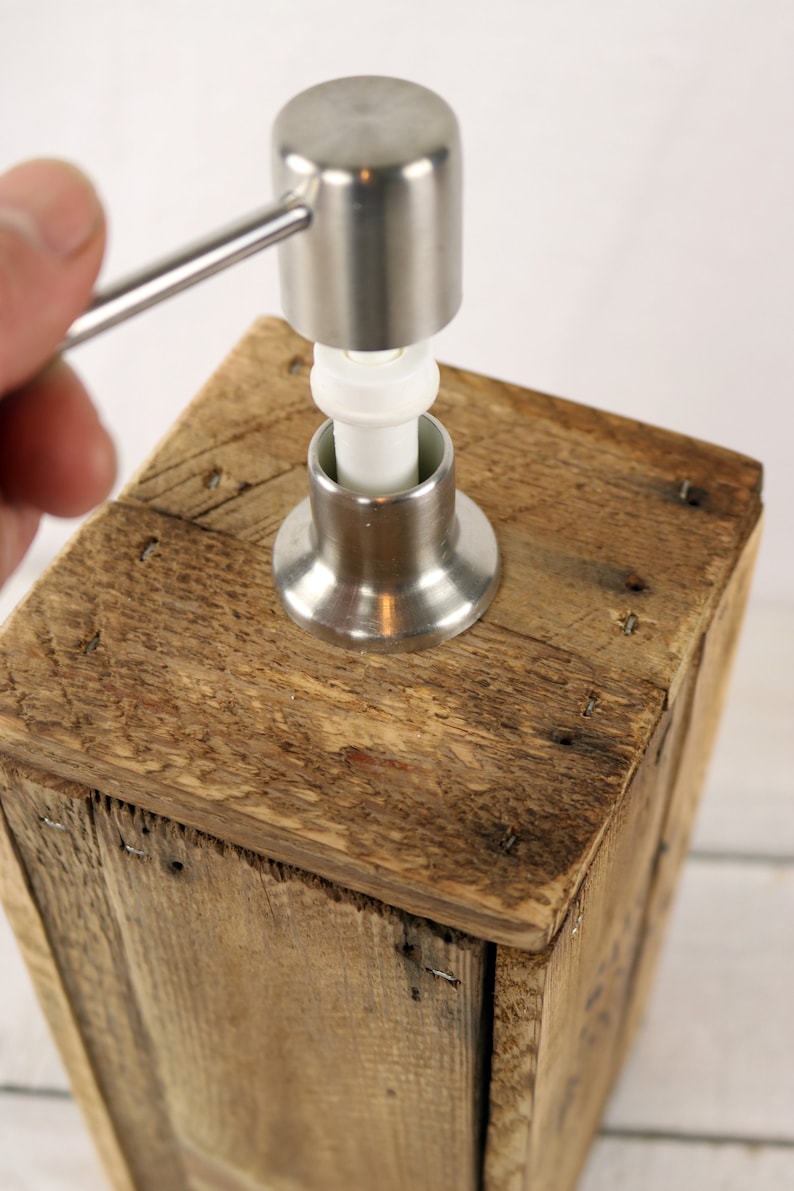 May include: A wooden soap dispenser with a stainless steel pump. The pump has a white plastic ring and a silver handle.