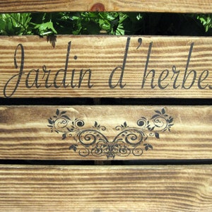 May include: A wooden sign with the text "Jardin d'herbes" in black lettering. The sign is made of weathered wood with a natural wood grain. A decorative floral design is below the text.