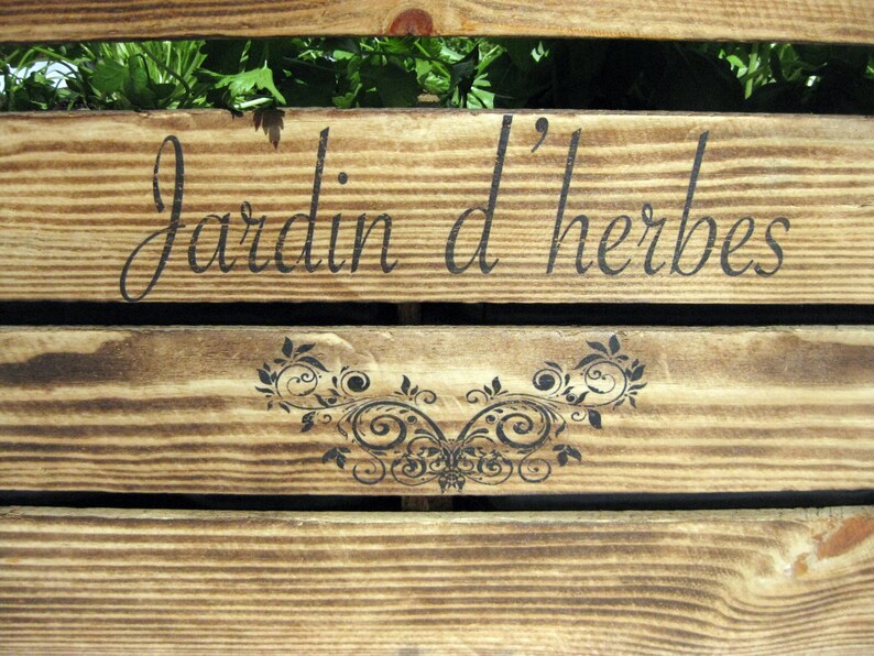 May include: A wooden sign with the text "Jardin d'herbes" in black lettering. The sign is made of weathered wood with a natural wood grain. A decorative floral design is below the text.