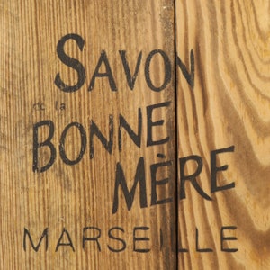 May include: A wooden sign with black lettering that reads "Savon la Bonne M&egrave;re Marseille".