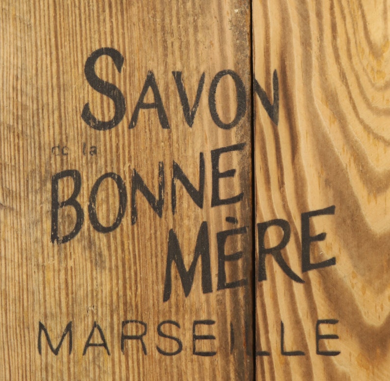 May include: A wooden sign with black lettering that reads "Savon la Bonne M&egrave;re Marseille".