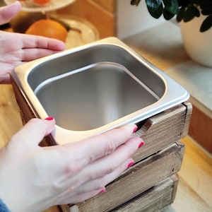 May include: A stainless steel food container with a wooden frame. The container is being held by a hand with red nail polish.