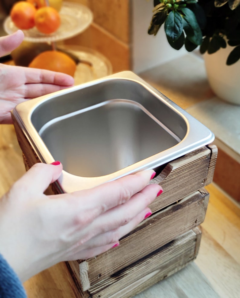 May include: A stainless steel food container with a wooden frame. The container is being held by a hand with red nail polish.