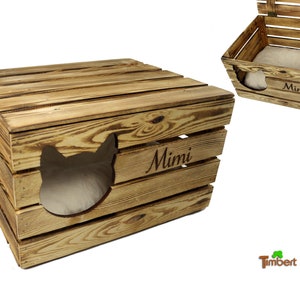 Op de afbeelding: Een houten kattenbed met een uitsparing in de vorm van een kattenkop en de naam "Mimi" gegraveerd op de voorkant. Het bed is gemaakt van natuurlijk hout met een rustieke afwerking.