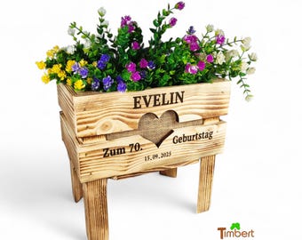 May include: A wooden crate planter with four legs, featuring a heart-shaped cutout and burlap backing. The front is engraved with the name EVELIN, the text Zum 70. GEBURTSTAG, and the date 15.09.2025. It is filled with yellow, purple, and white flowers.