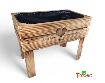 May include: A rectangular wooden planter box with four legs, featuring a dark brown stained finish and a black plastic liner. The front panel displays a heart-shaped cutout, burlap fabric backing, and the text Alles Liebe Mami 09.10.1964 zum Geburtstag.