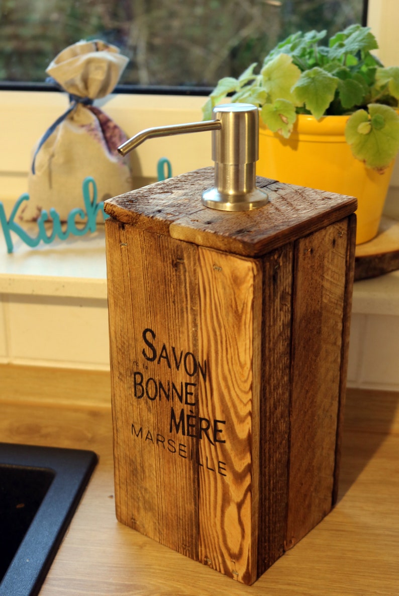 May include: A wooden soap dispenser with a stainless steel pump. The dispenser is labeled "Savon Bonne Mere Marseille".